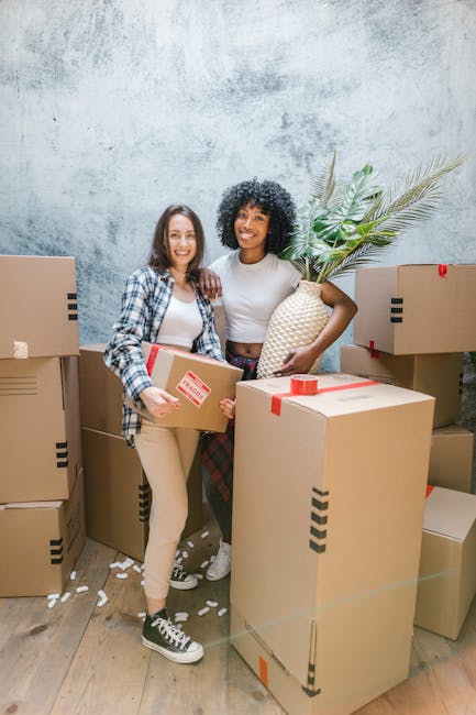 Two women standing inside a room filled with packed cardboard moving boxes, some sealed with red tape and others open with packing paper visible. One woman, on the left, is holding a small box and wearing a plaid shirt and beige pants, while the other, on the right, is holding a large decorative vase with a green plant and is dressed in a white top and checkered trousers. They are smiling and appear to be preparing for home relocation or furniture transport. The background shows a textured light grey wall, and the ground is covered with a wooden floor. The scene captures the loading process typical of professional removals, with the women engaged in packing or organizing items for a house move, exemplifying a typical moving and packing scenario supported by Bow Man and Van.