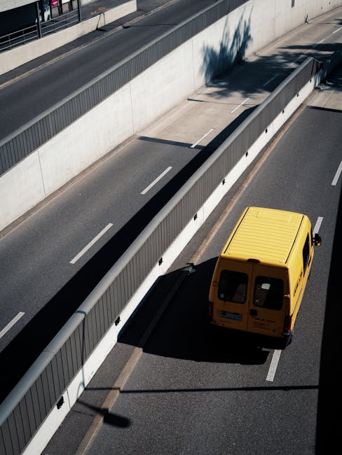 An aerial view of a yellow delivery van parked on the right side of a multi-lane road, with a tall concrete barrier and sound wall separating traffic lanes. The van is positioned adjacent to a loading zone, with a shadow cast by a tree visible on the roadway. The road surfaces are marked with white dashed lines indicating lane divisions, and a streetlamp is casting a shadow across the pavement. This scene illustrates an urban setting suitable for vehicle mobilization during home relocation or furniture transport tasks, with the vehicle prepared for loading or unloading near a designated area. The image emphasizes elements related to moving logistics, such as the parked van, nearby barriers, and the surrounding environment, aligning with professional removal services like those offered by Bow Man and Van.