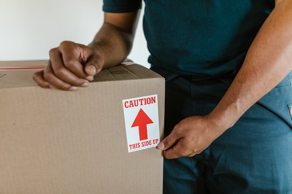 A close-up of a person wearing a dark green shirt and blue trousers, securing a cardboard box with a label that reads 'Caution This Side Up' and an upward arrow, indicating careful handling during a home relocation. The individual is gripping the box with both hands, one on each side, standing inside a property near an open doorway. The scene suggests the packing and moving process, emphasizing the importance of correct orientation for fragile or valuable items. Surrounding the person are additional moving materials, such as packing paper and plastic wrap, used for protection of furniture and household items during transportation. The focus on the handling label and the person's grip reflects the careful loading process at the start of furniture transport or house removals, with a van likely nearby for loading those packed boxes. The environment is well-lit, and the setting indicates a typical moving day, with commercial or professional removals services like Bow Man and Van assisting with relocation tasks near Victoria Park Bow.