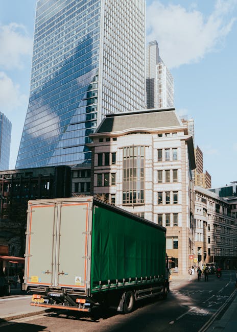 A large moving truck with a green cargo area is parked on a city street in front of modern high-rise buildings and older architectural structures. The truck, which appears to be involved in a home rel